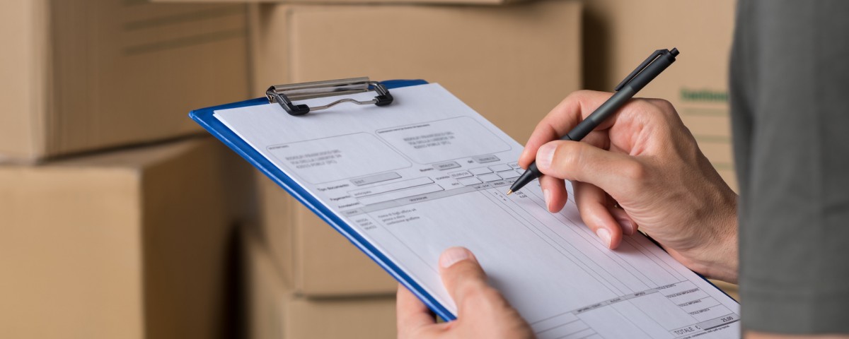 Warehouse manager signing dispatch sheet. Close up hand of manager checking delivery order. Young freight manager checking invoice with cardboard box parcel.